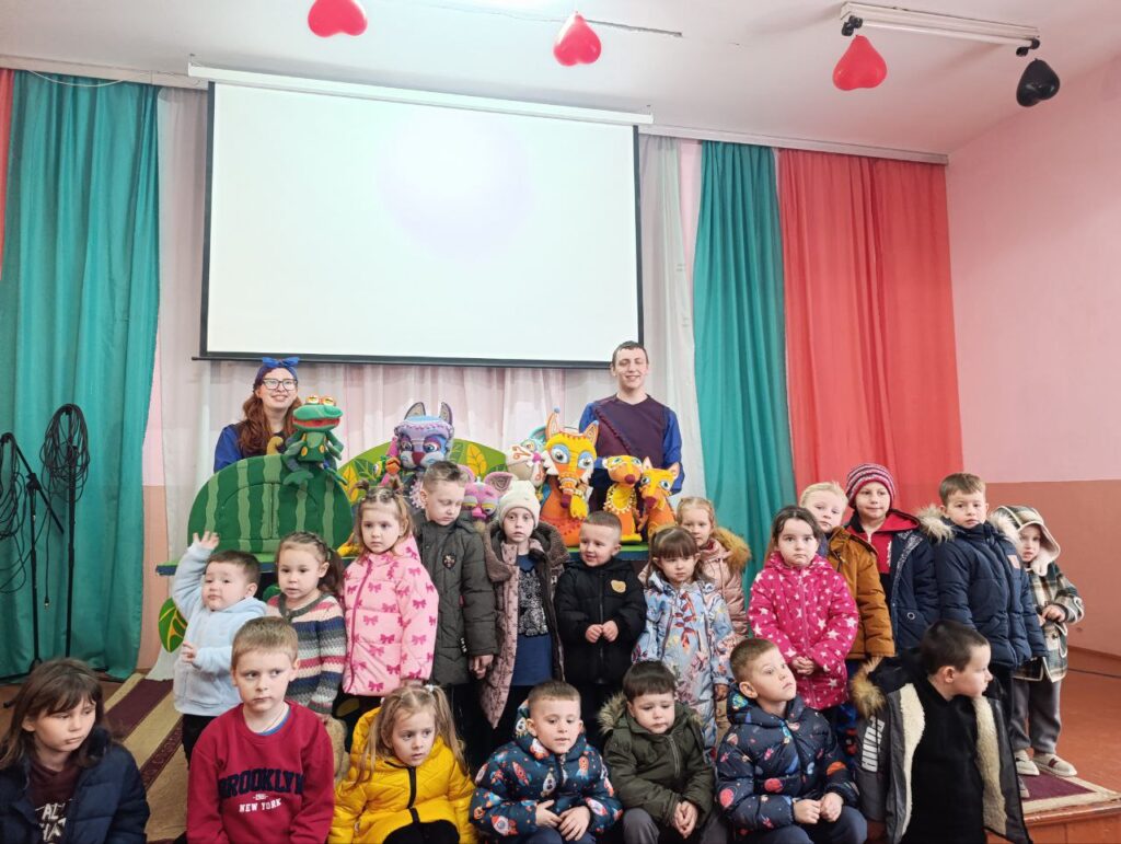 рупа маленьких дітей у зимовому одязі позує перед ляльковим театром. Позаду — двоє дорослих із ляльками: жабою, котом та іншими персонажами. Сцена прикрашена кольоровими завісами, над нею — великий екран.