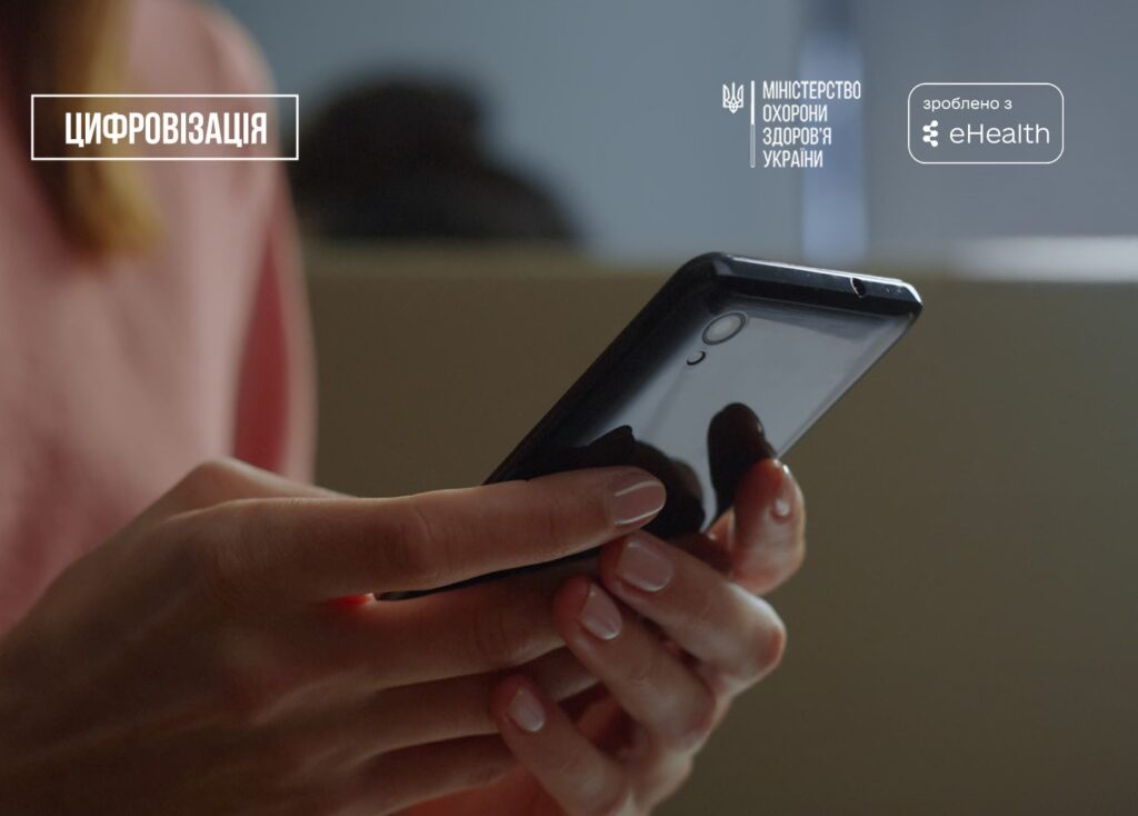 Руки людини у рожевій кофті тримають смартфон. На екрані зображення з логотипами та текстом про цифровізацію у сфері охорони здоров’я.