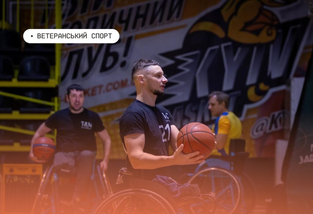 Група спортсменів у візках грає в баскетбол у спортивній залі; один гравець у чорній футболці з номером «21» тримає м’яч