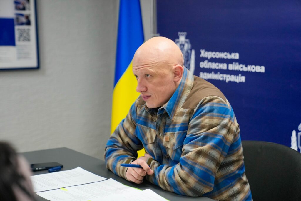 Чоловік у картатій сорочці сидить за столом із документами та ручкою в руках. Позаду синій банер із написом «Херсонська обласна військова адміністрація» та прапор України.