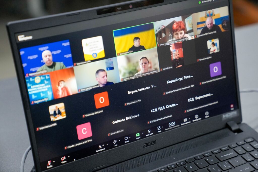 На екрані ноутбука — Zoom-конференція з багатьма учасниками. Деякі з камерами, інші з іконками чи іменами. У віконцях видно офіційні назви українською: «Херсонська ОВА», «Служба у справах дітей», «Бериславська РДА» тощо. 