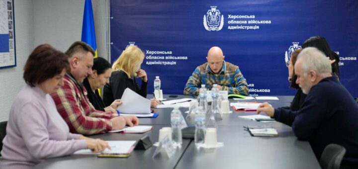 Дев’ятеро людей сидять за довгим столом із документами та пляшками води. Позаду синій банер із гербом та написом «Херсонська обласна військова адміністрація». Видно прапор України.