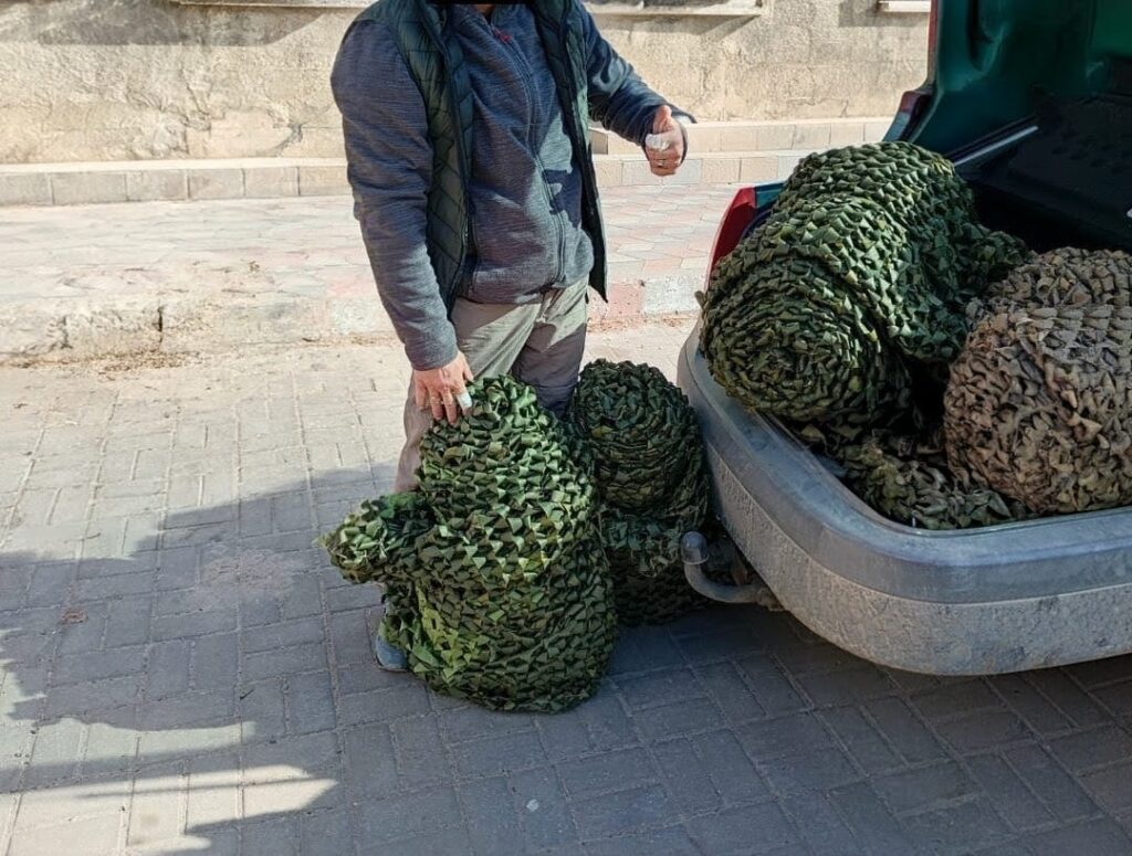 Людина у сірій куртці та бежевих штанах стоїть біля відкритого багажника автомобіля. Усередині — численні згорнуті камуфляжні сітки зеленого та коричневого кольору. На землі поруч — одна розгорнута сітка, яку тримає ця людина.