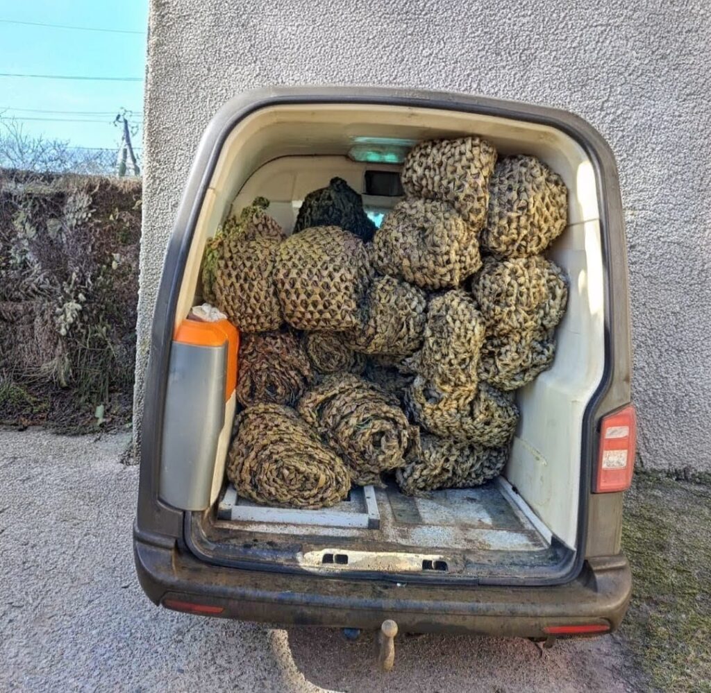 ідкриті задні двері фургона, всередині щільно складені рулони камуфляжних сіток у відтінках зеленого, коричневого та бежевого. Ліворуч — контейнер помаранчевого й сірого кольору. Фургон припаркований біля стіни, поруч видно кущі.