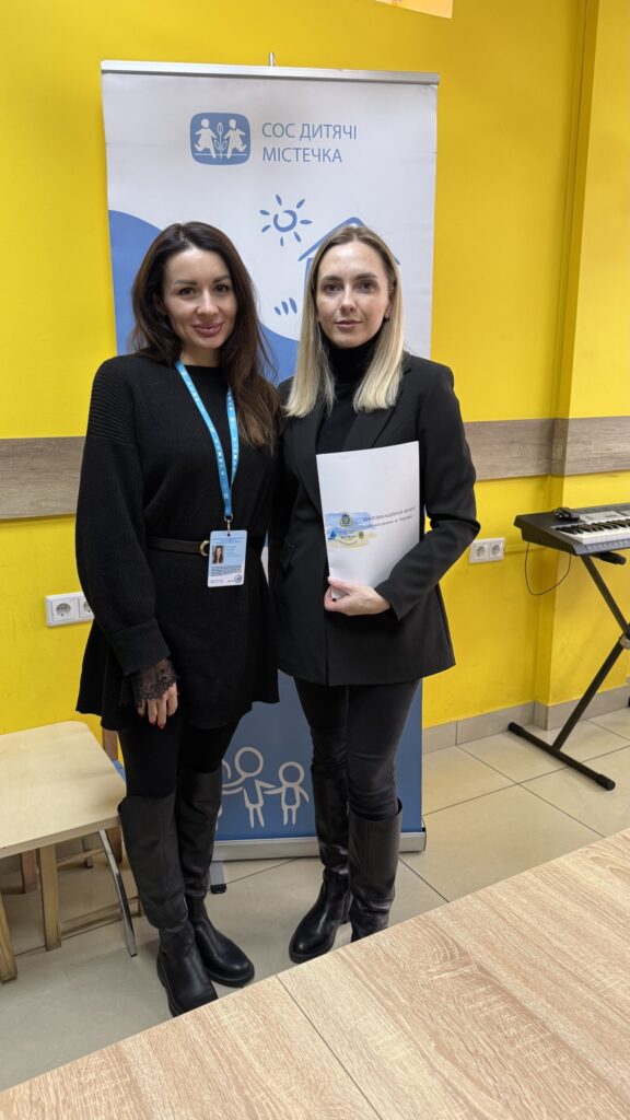 Двоє людей стоять у приміщенні перед банером із написом «SOS Дитячі містечка» та логотипом із дітьми, сонцем і будиночком. Ліворуч — жінка в чорному одязі з синім бейджем на ланцюжку. Праворуч — жінка в чорному жакеті тримає папку з написом «Харківський обласний центр» та логотипами. Позаду — жовта стіна, клавіатура на підставці та дерев’яні меблі.