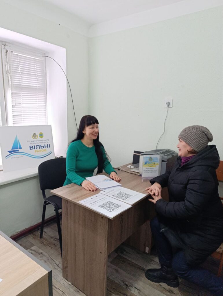 У невеликому офісі двоє людей сидять за столом один навпроти одного. Жінка з довгим темним волоссям у зеленому светрі працює за столом, перед нею документи. Навпроти — людина у зимовій куртці та в’язаній шапці. На столі лежать аркуші з QR-кодами та інформаційними надписами «Скануй для отримання допомоги». Є табличка з написом «Координаційний центр Чернівецької області» та «Вільні разом», поруч логотип із синім вітрильником та офіційними емблемами.