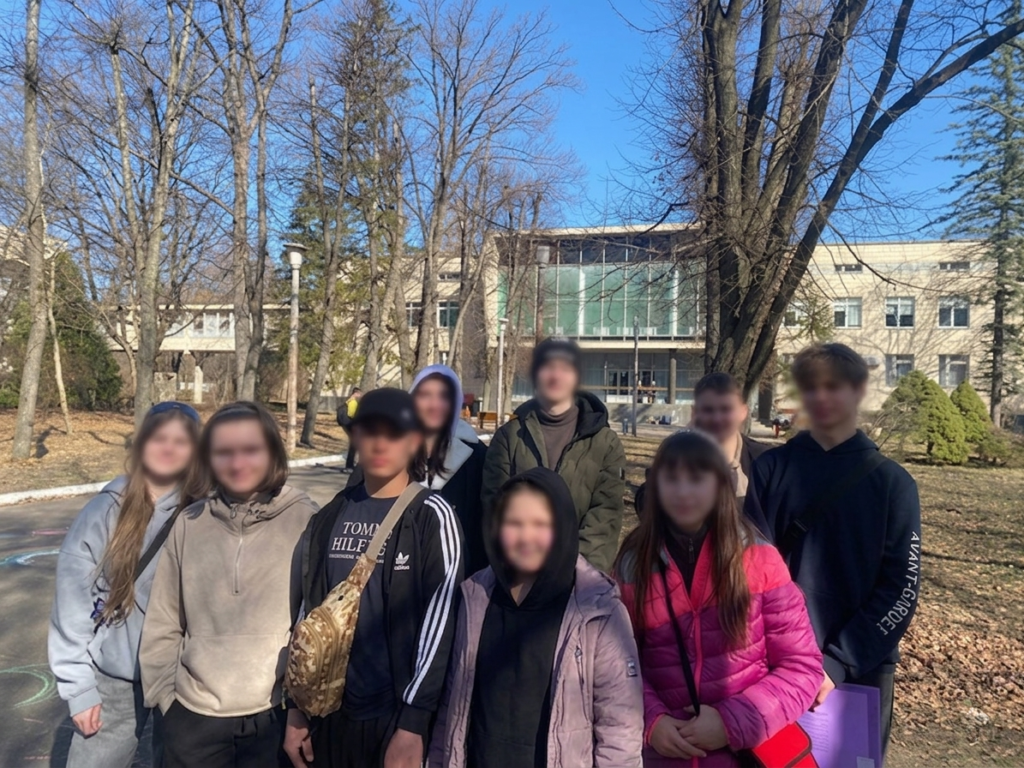 Дев’ятеро людей стоять надворі серед дерев без листя, сонячна погода. Усі в повсякденному одязі — куртки, худі, пальта. Позаду сучасна будівля зі скляними вікнами. На землі видно кольорові малюнки крейдою.