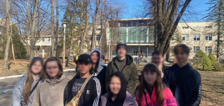 Дев’ятеро людей стоять надворі серед дерев без листя, сонячна погода. Усі в повсякденному одязі — куртки, худі, пальта. Позаду сучасна будівля зі скляними вікнами. На землі видно кольорові малюнки крейдою.