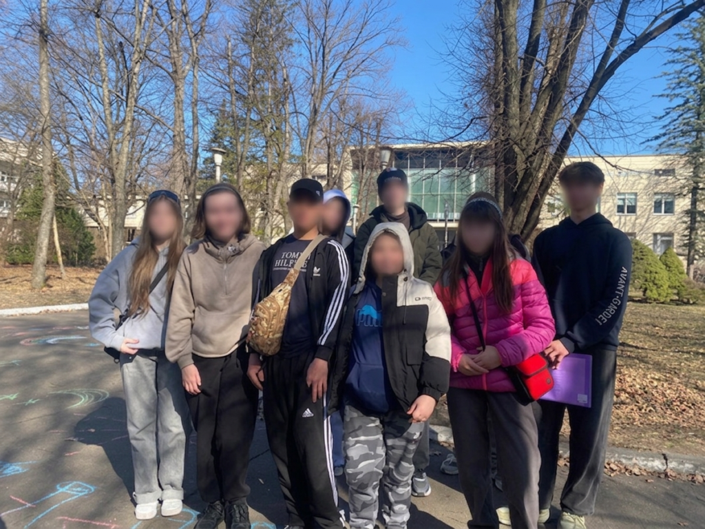 Вісім людей стоять на плитковому майданчику серед голих дерев. Одягнені у спортивні штани, куртки, худі. Дехто тримає речі — папку чи сумку. На землі — яскраві малюнки крейдою, що додають грайливості. Позаду — будівлі з великими вікнами.