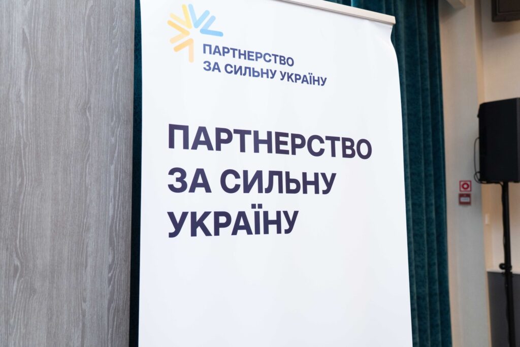 Вертикальний банер із кольоровим логотипом у вигляді променів синього, жовтого та помаранчевого кольорів. Текст: «Партнерство за сильну Україну». Банер стоїть у приміщенні біля дерев’яної стіни та темно-зелених завіс.