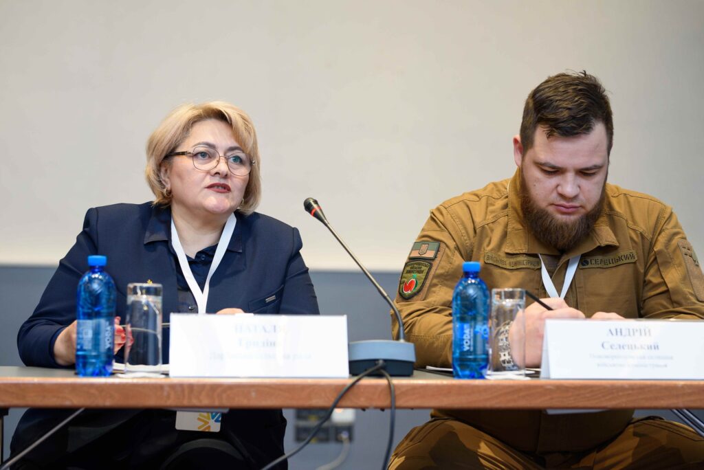 За столом сидять двоє учасників. Жінка в темно-синьому костюмі говорить у мікрофон, перед нею табличка «Наталія Гргайло, Державна служба якості». Чоловік у коричневій формі з військовими відзнаками пише нотатки, перед ним табличка «Андрій Селецький, Повноважний представник військової адміністрації».