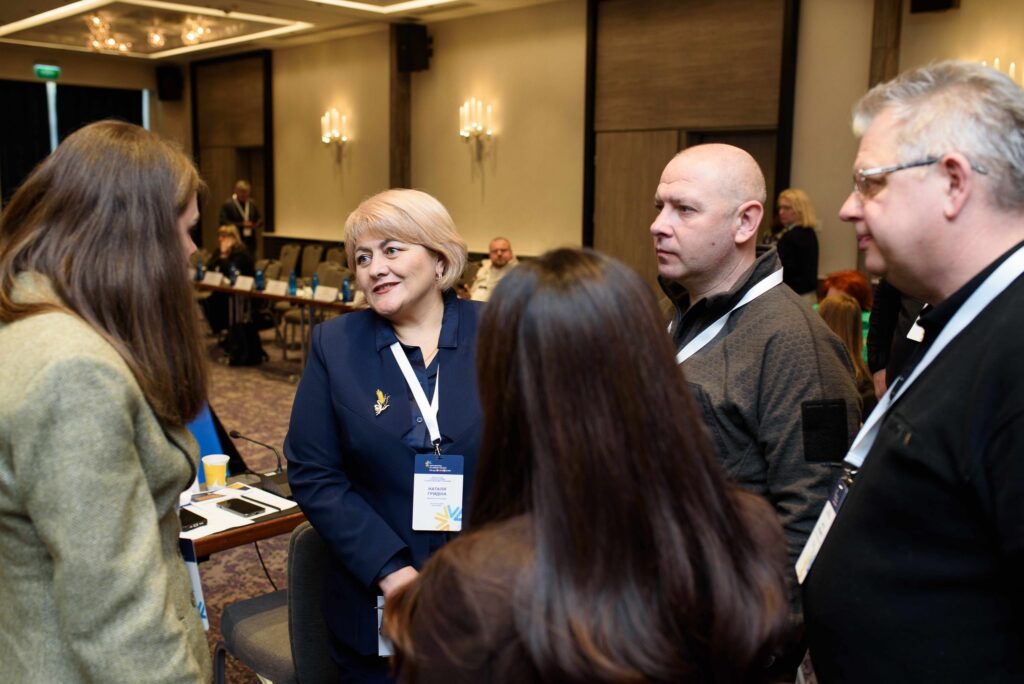 П’ятеро людей у бейджах розмовляють у залі з столами та учасниками. У центрі — жінка з бейджем «Natalia Hryniv».