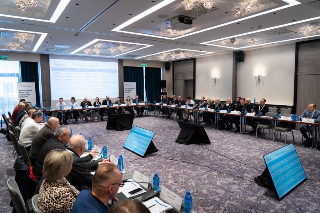 Сучасна конференц-зала з великим U-подібним столом, учасники з бейджами та документами. На екрані — слайд «Громади-учасники». По боках — банери «Partnership Fund for a Resilient Ukraine».