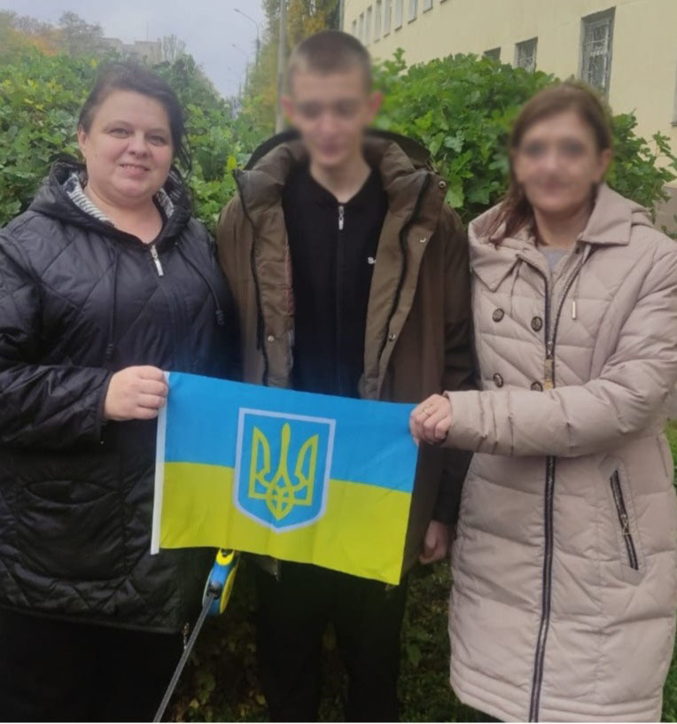 Троє людей стоять надворі перед зеленим живоплотом і будівлею. Усі в теплих куртках: чорній, коричневій та бежевій. В руках тримають маленький український прапор із синьо-жовтими смугами та тризубом посередині.