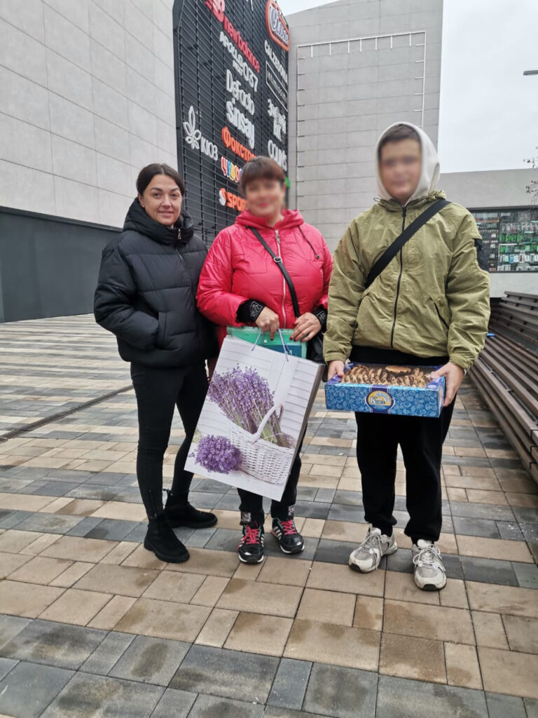 Троє людей стоять на плитковому майданчику перед сучасною будівлею. Ліворуч — чоловік у чорній куртці, посередині — жінка в червоній куртці з великою подарунковою сумкою з квітковим малюнком, праворуч — людина у світло-зеленій куртці з капюшоном тримає синю коробку з тістечками. Позаду — вертикальний знак із назвами магазинів 