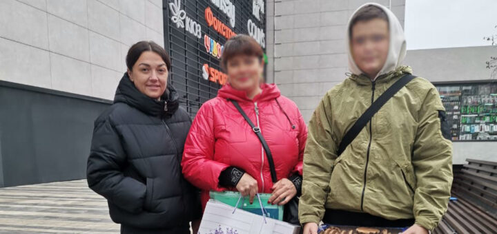 Троє людей стоять на плитковому майданчику перед сучасною будівлею. Ліворуч — чоловік у чорній куртці, посередині — жінка в червоній куртці з великою подарунковою сумкою з квітковим малюнком, праворуч — людина у світло-зеленій куртці з капюшоном тримає синю коробку з тістечками. Позаду — вертикальний знак із назвами магазинів