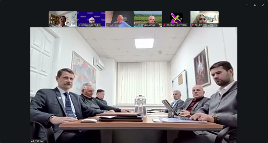 У центрі — семеро чоловіків сидять за прямокутним столом у світлому офісі. На столі документи, папки, графин із водою. На стінах — карта та картина. У верхній частині екрана — маленькі віконця інших учасників онлайн, з іменами «Ivo», «Eimantas», «Lado Uzunashvili» тощо.