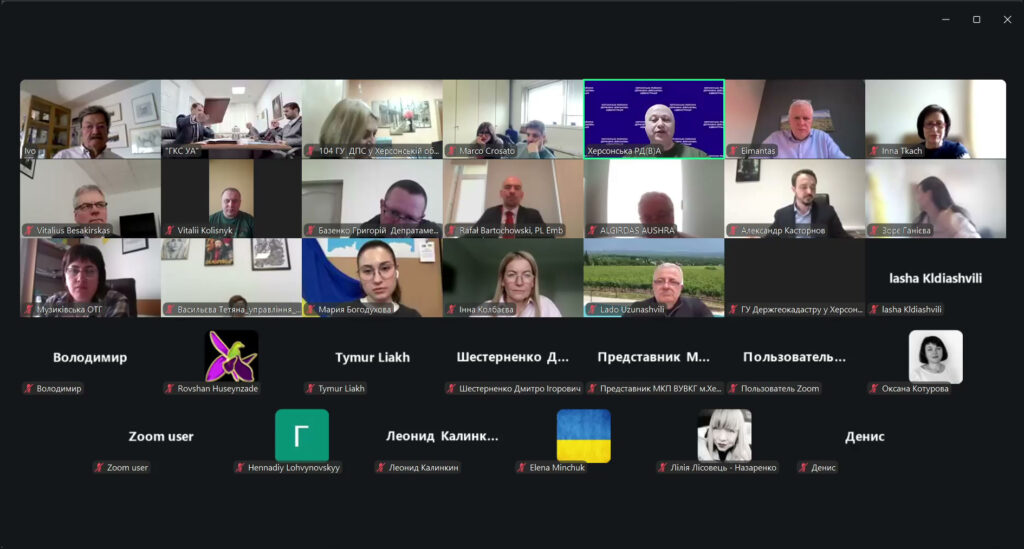 Скріншот відеоконференції у Zoom. Учасники відображені у сітці, більшість із вимкненими мікрофонами. Видно офіційні фони, прапори та формальний одяг. Імена учасників написані кирилицею й латиницею, що свідчить про міжнародну участь. 