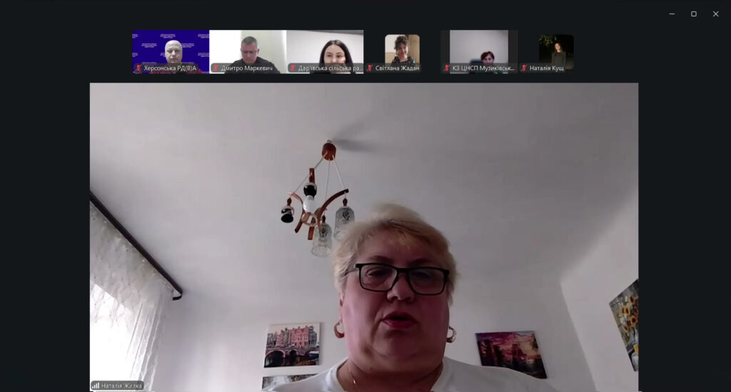 Жінка у білому одязі та окулярах сидить у світлій кімнаті з картинами на стіні. У верхній частині — маленькі віконця інших учасників із назвами