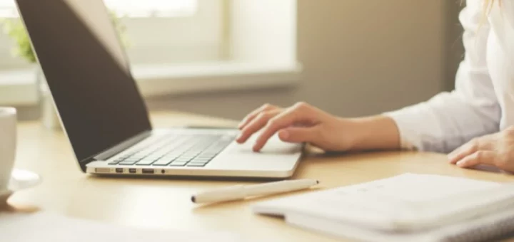 Робоче місце з ноутбуком, рука людини на клавіатурі, поруч ручка, блокнот і чашка, на тлі — вікно з денним світлом