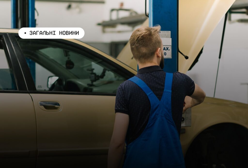 Механік у синьому комбінезоні працює біля автомобіля з відкритим капотом у майстерні. Поруч — синій гідравлічний підйомник.