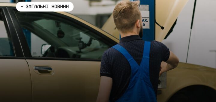 Механік у синьому комбінезоні працює біля автомобіля з відкритим капотом у майстерні. Поруч — синій гідравлічний підйомник.