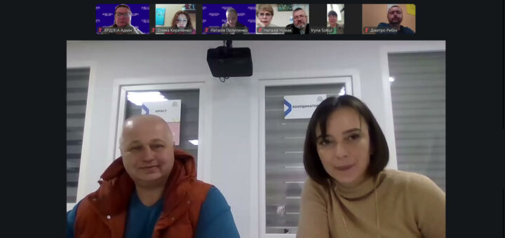 У центрі — двоє людей сидять поруч у світлій кімнаті з вікнами позаду. Ліворуч — чоловік у синій сорочці та помаранчевому жилеті, праворуч — жінка у бежевому светрі. На вікнах видно таблички «Координатор» та «Юрист». У верхній частині — маленькі віконця інших учасників із іменами