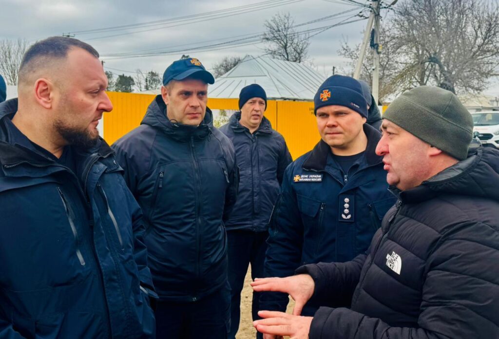 П’ятеро чоловіків стоять надворі й ведуть серйозну розмову. Чотири — у темно-синіх формах із нашивками «ДСНС України» та «Рятівники». П’ятий — у чорній куртці з логотипом «The North Face» і зеленій шапці, активно жестикулює. На тлі — жовта стіна чи контейнер, металевий дах, дерева без листя та дроти під хмарним небом. 