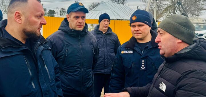 П’ятеро чоловіків стоять надворі й ведуть серйозну розмову. Чотири — у темно-синіх формах із нашивками «ДСНС України» та «Рятівники». П’ятий — у чорній куртці з логотипом «The North Face» і зеленій шапці, активно жестикулює. На тлі — жовта стіна чи контейнер, металевий дах, дерева без листя та дроти під хмарним небом.