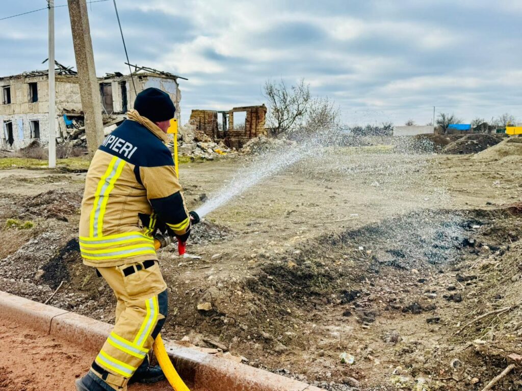 Пожежник у повному захисному спорядженні тримає шланг і поливає водою ділянку обгорілої землі. На спині куртки напис «POMPIERI». На тлі — пошкоджені та частково зруйновані будівлі, похмуре небо.