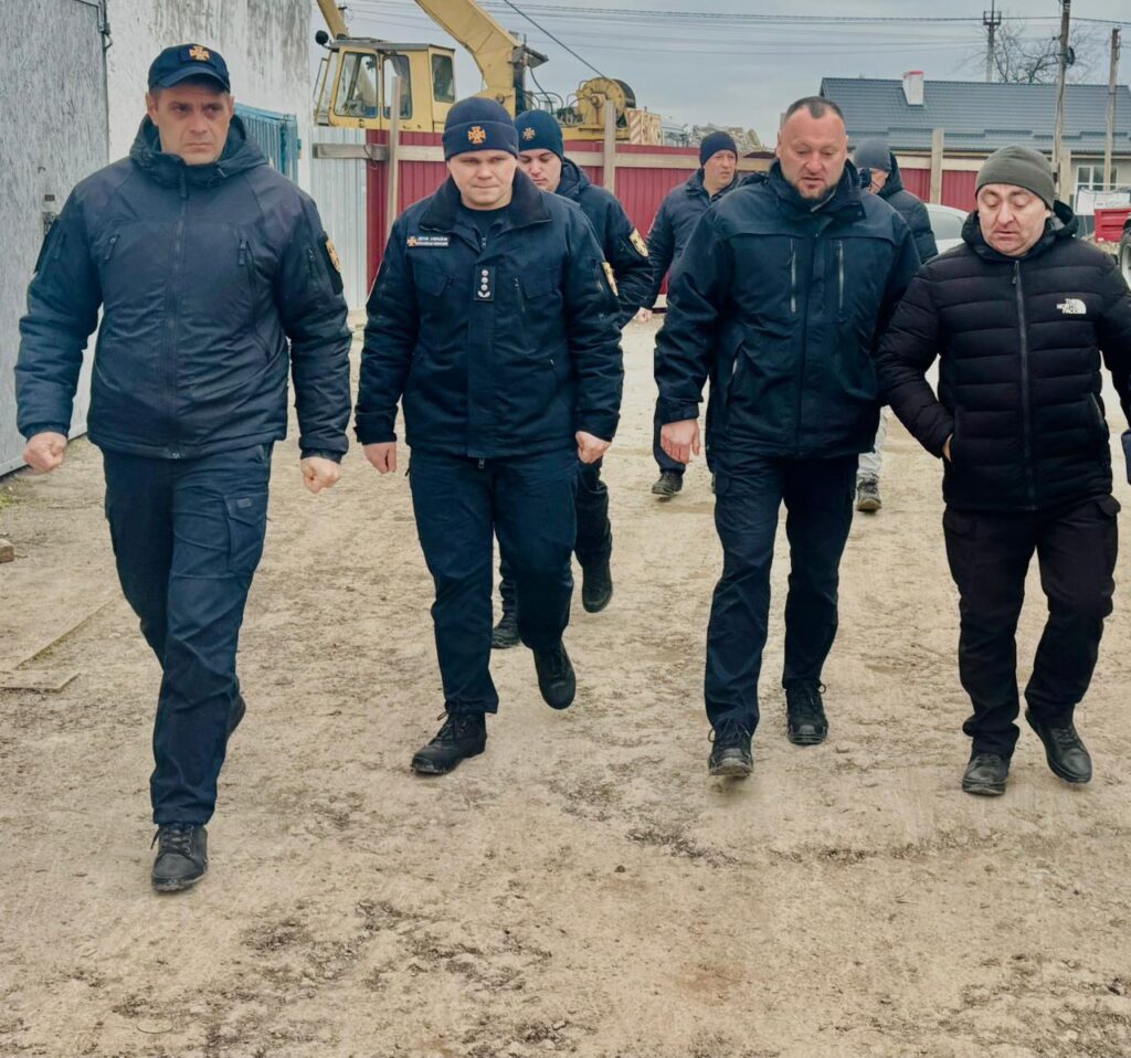 П’ятеро чоловіків у темних формах і куртках йдуть ґрунтовою стежкою. Деякі носять шапки чи бейсболки. На формі — нашивки та відзнаки, що свідчать про належність до силових чи охоронних структур. На тлі — екскаватор, кран, червоний паркан і житлові будинки. 