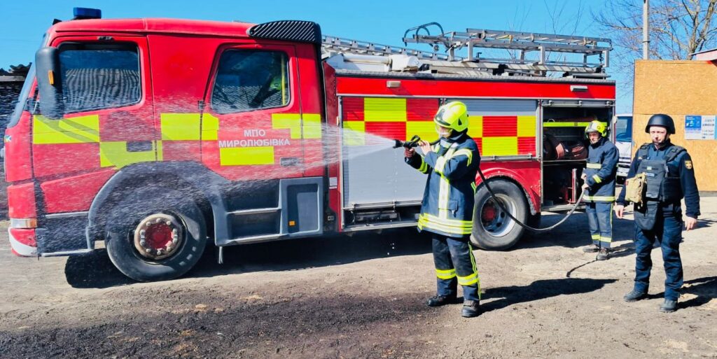 Червона пожежна машина з жовтими світловідбивними смугами стоїть на ґрунтовій поверхні. На боці напис «МПО Миролюбівка». Двоє пожежників у повному захисному спорядженні з касками працюють зі шлангом: один тримає та поливає водою, інший контролює підключення. Поруч третя людина у чорній формі з каскою та тактичним жилетом спостерігає. 