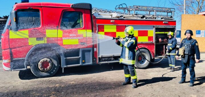 Червона пожежна машина з жовтими світловідбивними смугами стоїть на ґрунтовій поверхні. На боці напис «МПО Миролюбівка». Двоє пожежників у повному захисному спорядженні з касками працюють зі шлангом: один тримає та поливає водою, інший контролює підключення. Поруч третя людина у чорній формі з каскою та тактичним жилетом спостерігає.