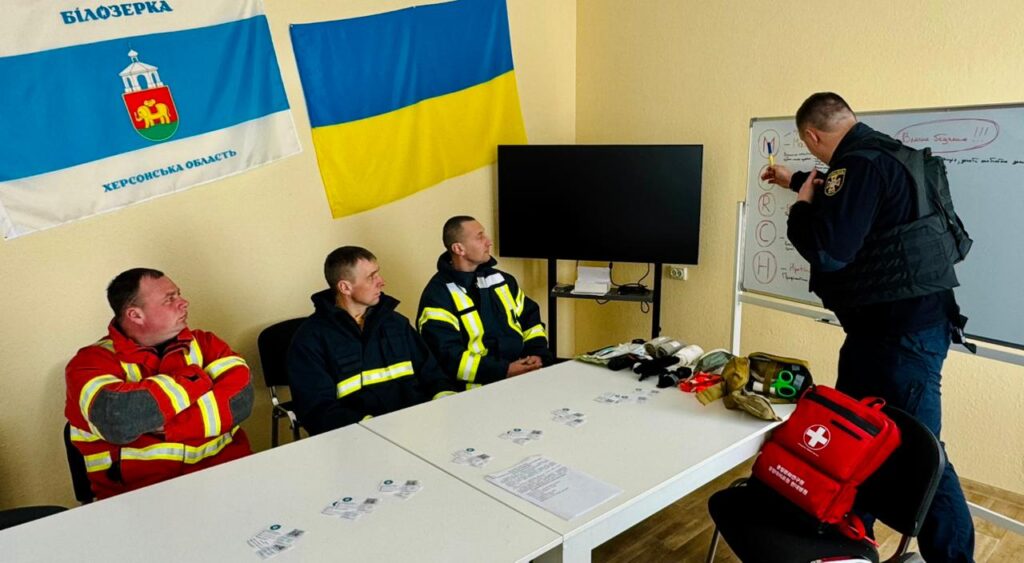 У кімнаті четверо людей: троє сидять у формі з відбиваючими смугами, один стоїть біля дошки й пояснює. На дошці написано «MARCH» вертикально. На столі — медичні матеріали: бинти, ножиці, аптечка. Позаду прапор України та прапор «Білозерка, Херсонська область». 