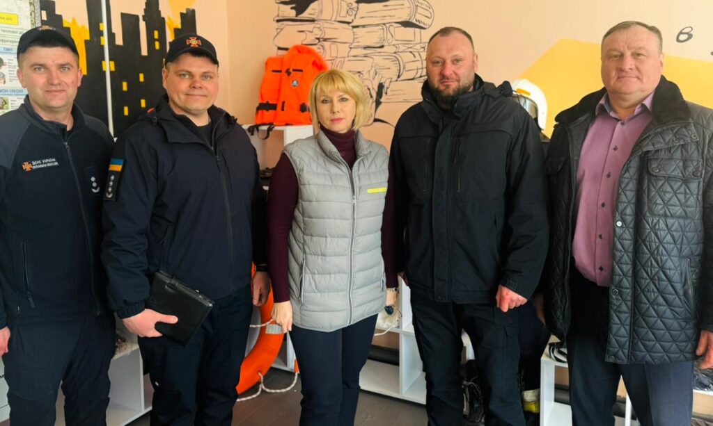 П’ятеро людей стоять перед стіною з муралом книжок і міста. Троє у темних формах із нашивками «ДСНС України» та «Миколаївська область». У центрі жінка у світло-сірій жилетці, праворуч двоє чоловіків у цивільному. Позаду — рятувальні жилети й круг. 
