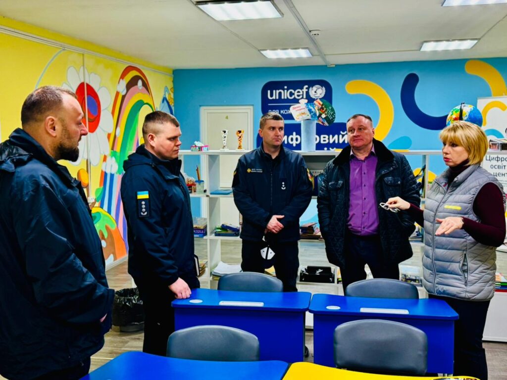 П’ятеро дорослих стоять у класі з барвистими малюнками на стінах — веселка, квіти, мультяшні елементи. Позаду полиці з книжками й трофеями. На синій стіні логотип UNICEF та напис «Цифровий освітній центр». 