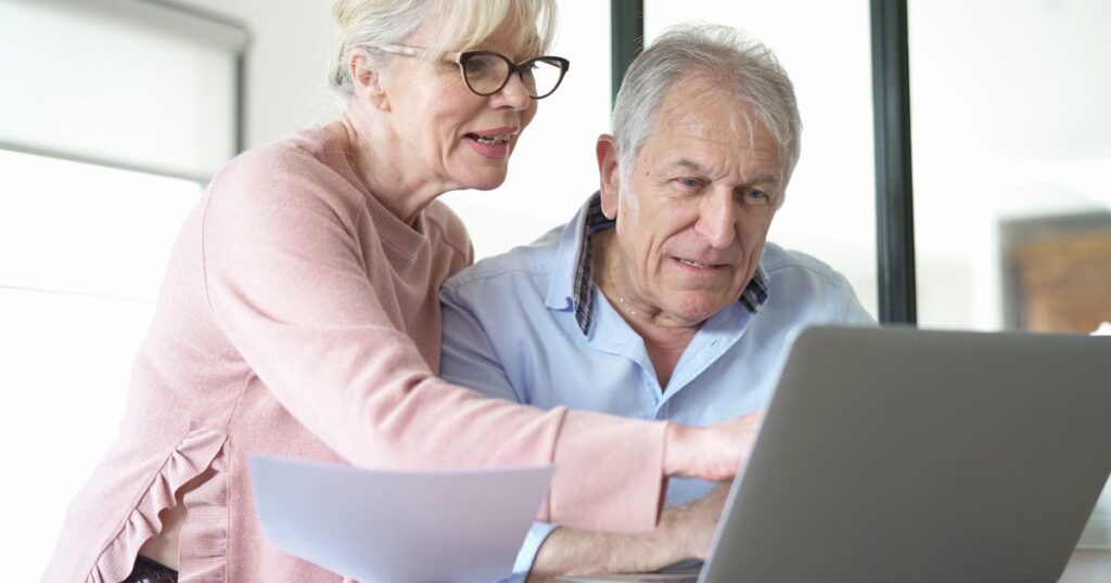 Двоє літніх людей сидять за столом, дивляться на екран ноутбука, один тримає аркуш паперу