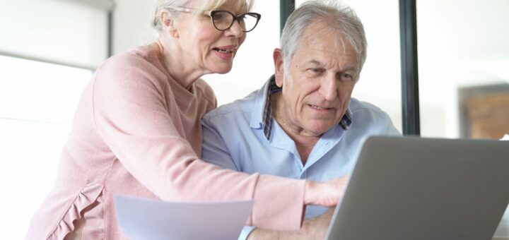 Двоє літніх людей сидять за столом, дивляться на екран ноутбука, один тримає аркуш паперу