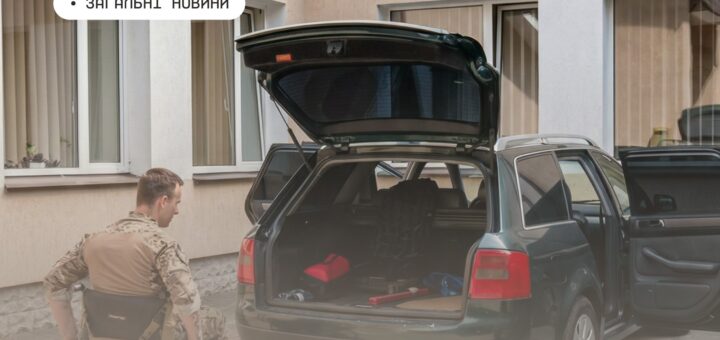 Людина у візку біля відкритого багажника зеленого універсала, поруч інструменти та речі