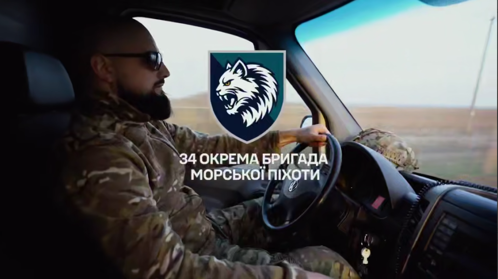 Військовий у камуфляжі та окулярах керує автомобілем, на зображенні емблема вовка й текст про 34-ту окрему бригаду морської піхоти