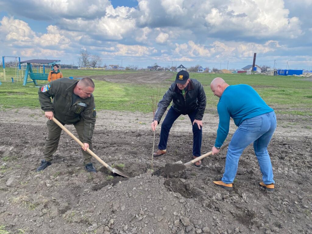 Троє людей працюють на відкритому ґрунті, саджаючи маленьке деревце. Двоє тримають лопати, один ставить саджанець. Позаду — поле, дитячий майданчик і будівлі.