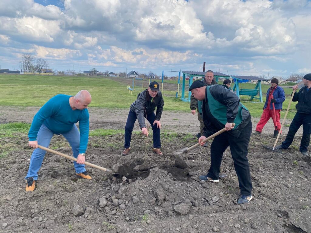 На ділянці землі кілька людей із лопатами копають чи перевертають ґрунт. Інші стоять поруч із інструментами. Позаду — поле й дитячі споруди.
