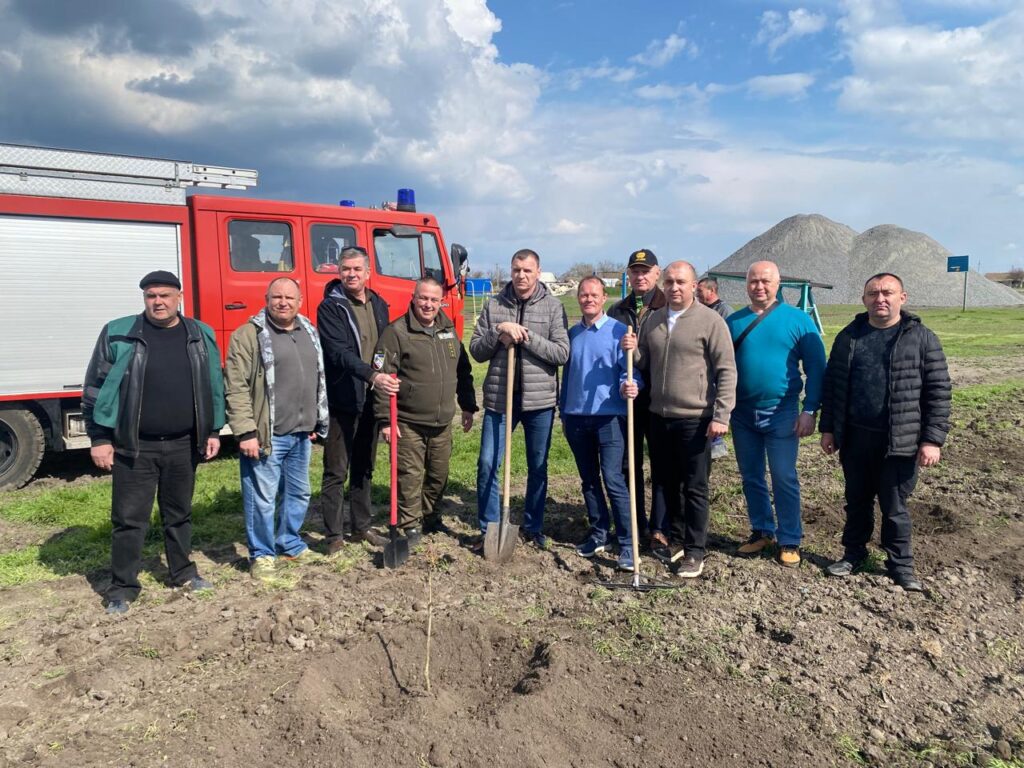 Одинадцять людей стоять на ділянці землі, де вже посаджено саджанець. У руках — лопати й граблі. Позаду — червона пожежна машина та купи гравію