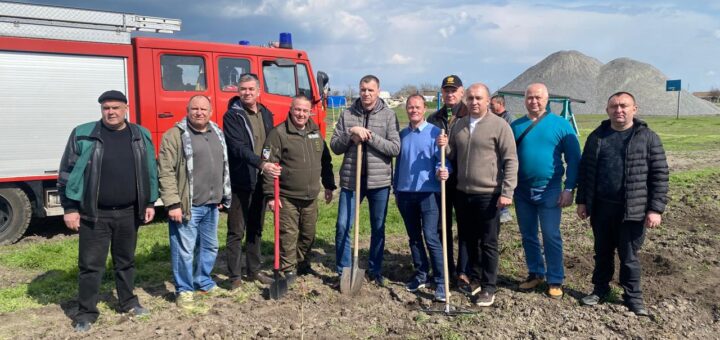 Одинадцять людей стоять на ділянці землі, де вже посаджено саджанець. У руках — лопати й граблі. Позаду — червона пожежна машина та купи гравію