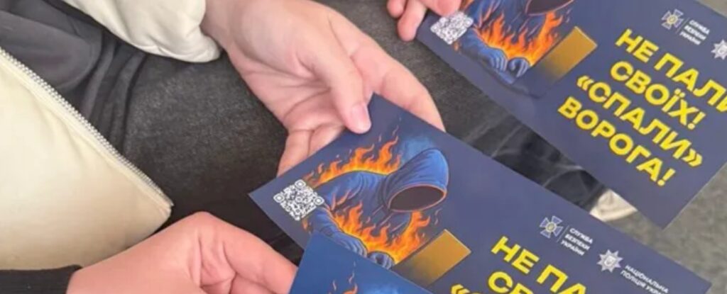 Дві руки тримають однакові листівки з темним фоном: постать у капюшоні перед ноутбуком у полум’ї, напис “Не пали своїх! ‘Спали’ ворога!”, логотипи СБУ та Національної поліції України