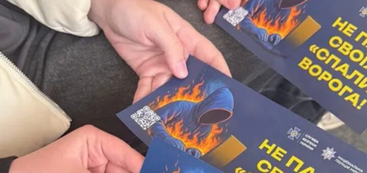 Дві руки тримають однакові листівки з темним фоном: постать у капюшоні перед ноутбуком у полум’ї, напис “Не пали своїх! ‘Спали’ ворога!”, логотипи СБУ та Національної поліції України