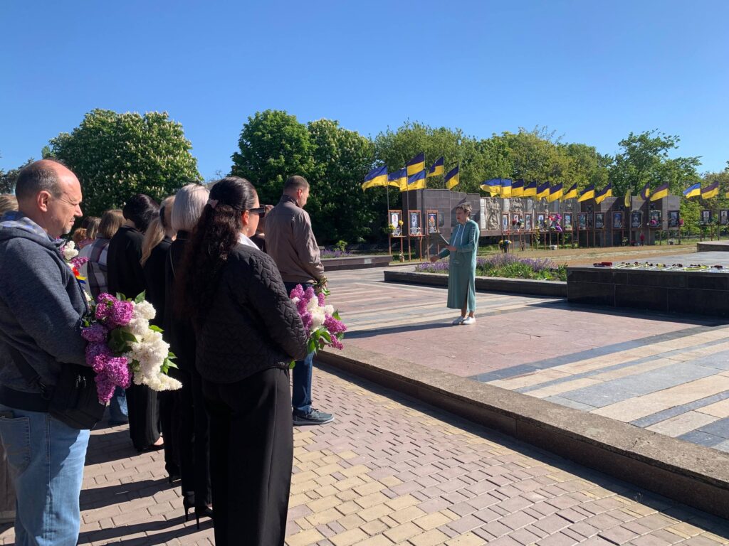 Група людей із бузком у руках стоїть перед стіною з фотографіями та прапорами. Попереду — ведуча у зеленому вбранні.