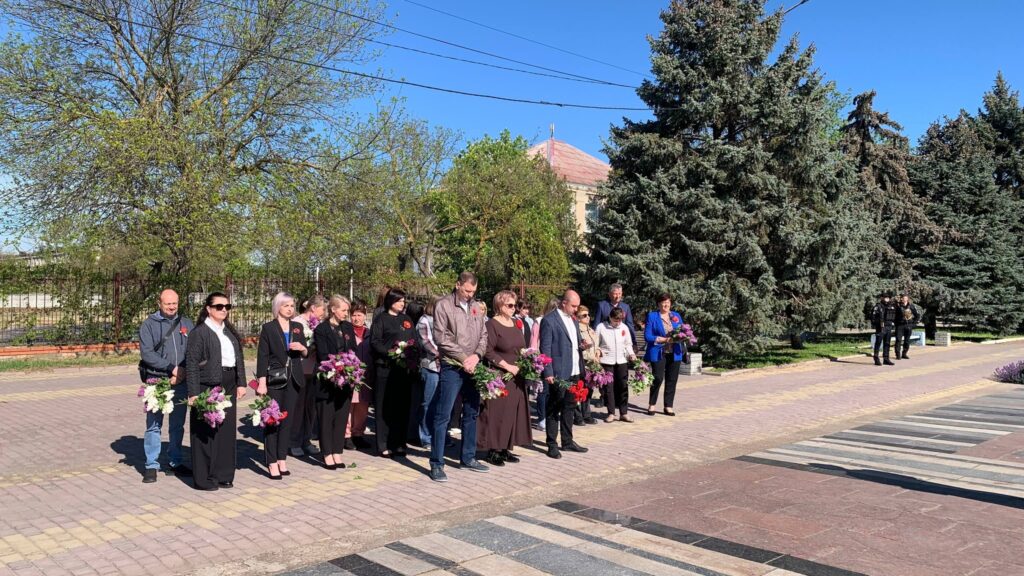 Люди з квітами стоять на алеї перед пам’ятником. Атмосфера — спільна пам’ятна церемонія.