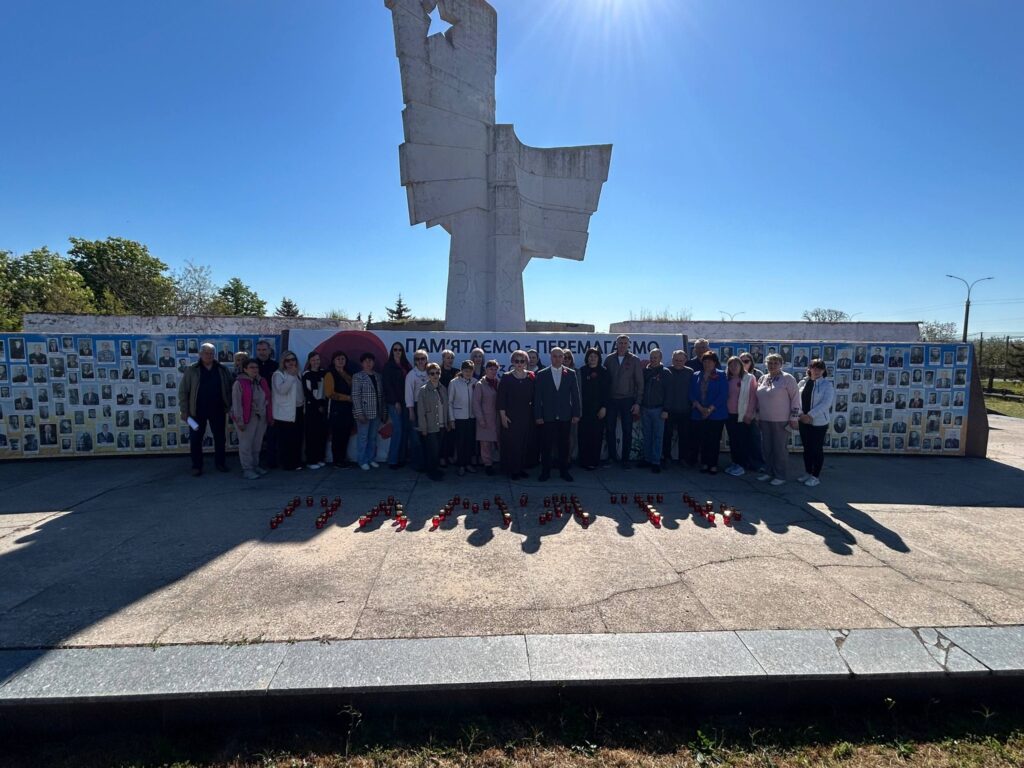 Група людей біля світлого пам’ятника. На стіні позаду — багато фотографій і банер «Пам’ятаємо — перемагаємо». На землі свічки викладають слово «Пам’ять». 