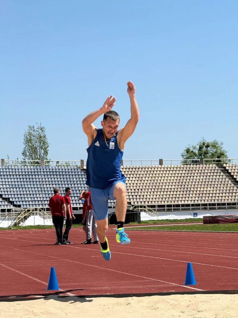 Спортсмен у синій формі виконує стрибок у довжину над піском. На доріжці — сині конуси, позаду люди й трибуни. 